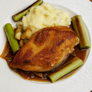 Pan-seared chicken breast steak served with sweet soy–maple onion sauce, mashed potatoes, garlic cloves, and spring onion pieces on a white plate.