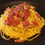 A plate of bacon garlic oil pasta served on a black dish, topped with crispy bacon pieces, sautéed garlic slices, and freshly ground black pepper.