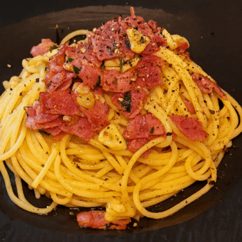 A plate of bacon garlic oil pasta served on a black dish, topped with crispy bacon pieces, sautéed garlic slices, and freshly ground black pepper.