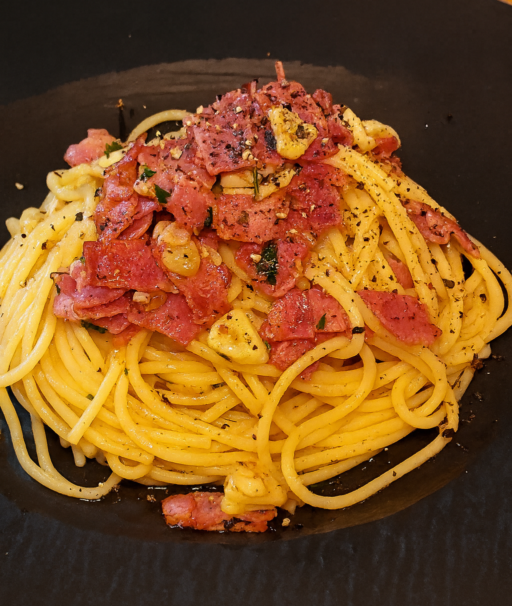 A plate of bacon garlic oil pasta served on a black dish, topped with crispy bacon pieces, sautéed garlic slices, and freshly ground black pepper.