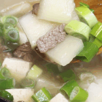 A ladle of Korean beef radish soup with brisket, tofu, radish cubes, and sliced green onions lifted from a pot.