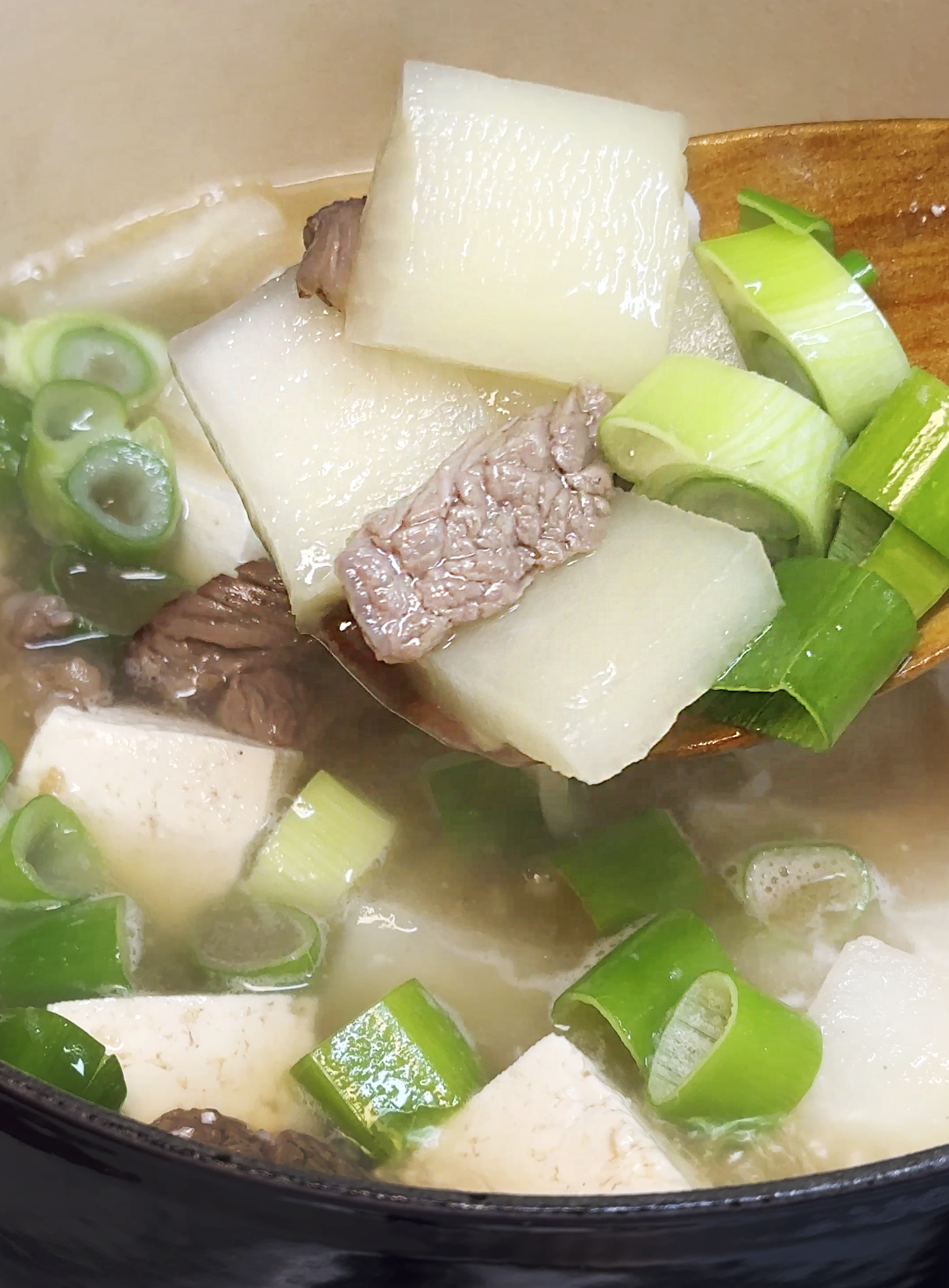 A ladle of Korean beef radish soup with brisket, tofu, radish cubes, and sliced green onions lifted from a pot.