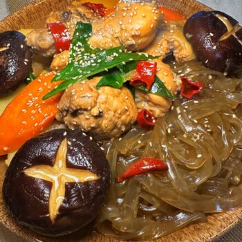 A dish of Jjimdak, or Korean braised chicken, served on a brown plate. It includes chicken pieces, glass noodles (japchae), large carrots, potatoes, and whole shiitake mushrooms garnished with sesame seeds and green onions.