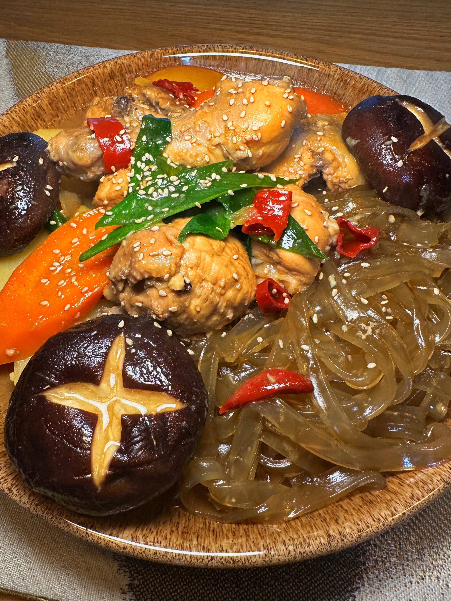 A dish of Jjimdak, or Korean braised chicken, served on a brown plate. It includes chicken pieces, glass noodles (japchae), large carrots, potatoes, and whole shiitake mushrooms garnished with sesame seeds and green onions.