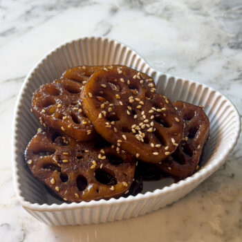 Heart-shaped dish filled with glossy Korean braised lotus root (yeongeun jorim) topped with sesame seeds.