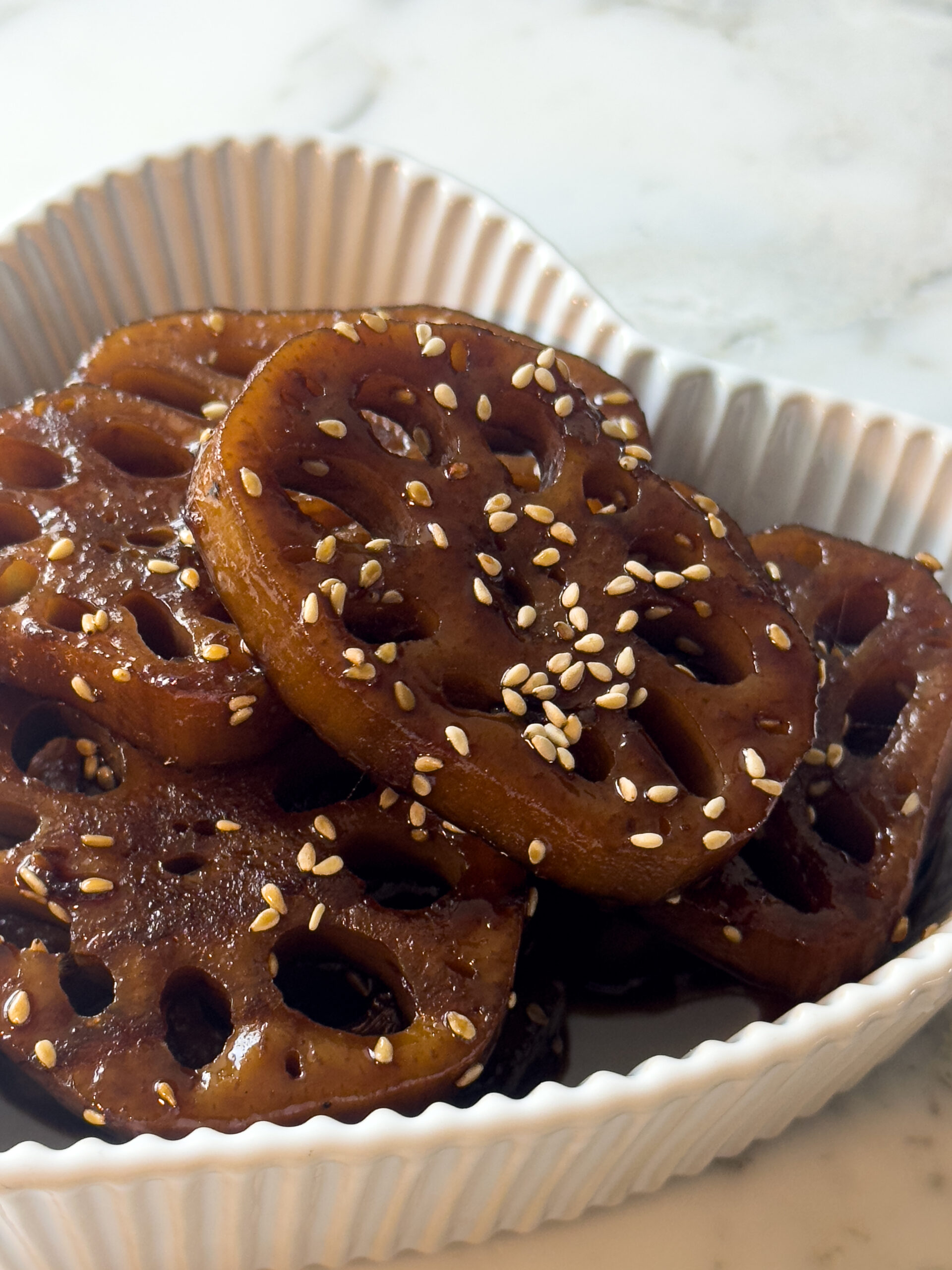 Close-up of glossy Korean braised lotus root (yeongeun jorim) topped with toasted sesame seeds in a white heart-shaped dish.